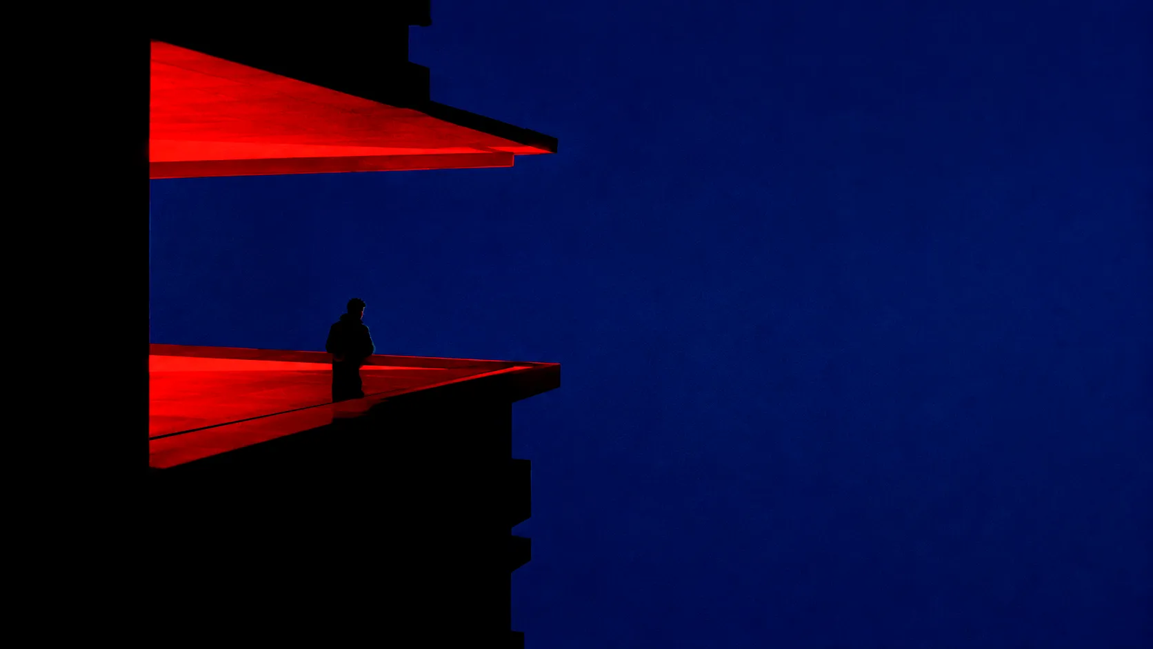 Un hombre en sombras, observando la inmensidad asomado a un balcón de un edificio en alturas.